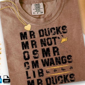 May include: A brown Comfort Colors t-shirt with the text "MR DUCKS MR NOT OSMR GM WANGS LIB MR DUCKS" in black. A gold heart pendant necklace and a beige hat are also visible. The shirt has a distressed look.