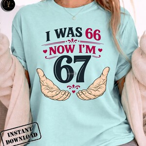 Puede incluir: Camiseta azul claro con el texto "I WAS 66 NOW I'M 67" en negro y rojo. El n&uacute;mero 67 es grande y centrado, con dos manos a cada lado. La camiseta tiene un gr&aacute;fico de "Descarga instant&aacute;nea".