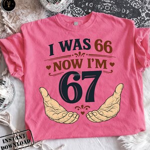 Puede incluir: Camiseta rosa con el texto "I WAS 66 NOW I'M 67" en azul oscuro y marr&oacute;n. El n&uacute;mero 67 est&aacute; en una fuente grande y estilizada, con dos manos ilustradas debajo. Un divertido regalo de cumplea&ntilde;os.