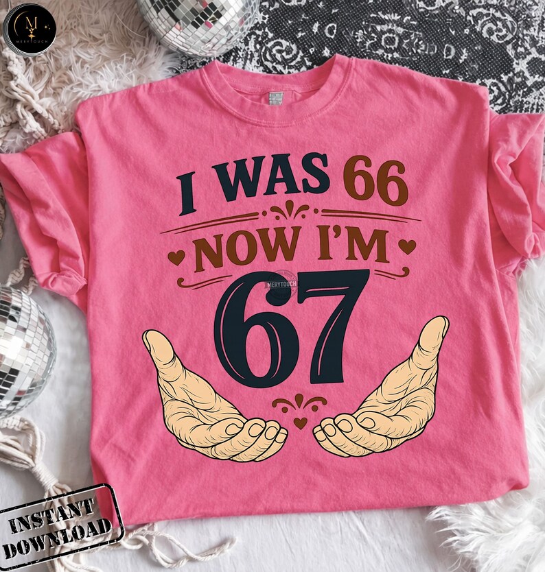 Puede incluir: Camiseta rosa con el texto "I WAS 66 NOW I'M 67" en azul oscuro y marr&oacute;n. El n&uacute;mero 67 est&aacute; en una fuente grande y estilizada, con dos manos ilustradas debajo. Un divertido regalo de cumplea&ntilde;os.