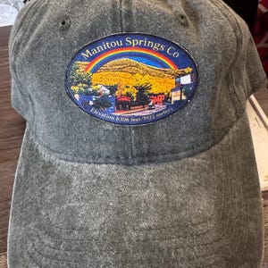 May include: A charcoal grey baseball cap featuring an oval embroidered patch on the front. The patch depicts a mountain landscape under a rainbow with the text Manitou Springs Co and Elevation 6306 feet / 1922 meters printed on the lower edge.