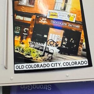 May include: A rectangular sticker featuring a photograph of the Rocky Mountain Chocolate Factory in Old Colorado City, Colorado. The image shows the store's exterior with text that reads "CHOCOLATE FACTORY" and "OLD COLORADO CITY, COLORADO."