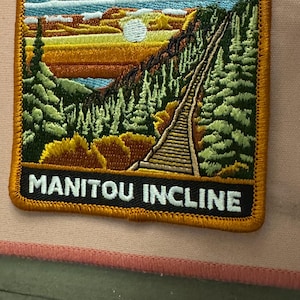 May include: Embroidered patch featuring a scenic landscape with a mountain incline, trees, and a sunset. The patch has a gold border and the text "MANITOU INCLINE" in white letters on a black background.