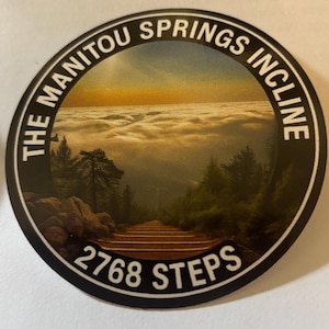 May include: Circular sticker featuring a scenic view of the Manitou Springs Incline, with a wooden staircase leading up through a forest towards a sea of clouds. The text "THE MANITOU SPRINGS INCLINE" and "2768 STEPS" are displayed.