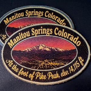 May include: Two oval embroidered patches featuring a sunset landscape of Pikes Peak. The text reads Manitou Springs Colorado and At the foot of Pike Peak, elev. 14,115 ft. The design includes a mountain range with snow and a city view at night.