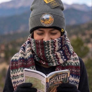 May include: A charcoal gray knit beanie with a Firm Grip tag and a circular patch featuring a mountain landscape. A thick, multi-colored patterned scarf in shades of burgundy, navy, and cream is wrapped around the neck. A small guidebook titled Manitou Springs Incline 2768 is held open, displaying a map and text.