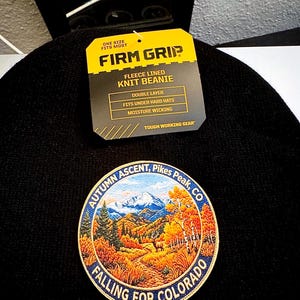 May include: A black knit beanie with a yellow product tag reading FIRM GRIP FLEECE LINED KNIT BEANIE. A circular patch features a colorful mountain landscape with an elk, text reading AUTUMN ASCENT, Pikes Peak, CO and FALLING FOR COLORADO.