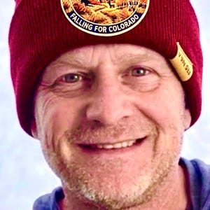 May include: A deep red knit beanie featuring a circular patch with a mountain landscape illustration. The patch text reads AUTUMN ASCENT, Pikes Peak, CO and FALLING FOR COLORADO. A small tan fabric tag is attached to the side of the ribbed cuff.