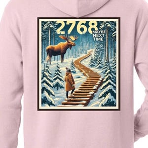 May include: A light pink hooded sweatshirt featuring a rectangular graphic on the back. The print depicts a snowy forest with a moose, a person walking up wooden stairs, and the text 2768 MAYBE NEXT TIME in a bold, dark font at the top.