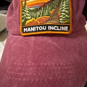 May include: A dusty rose baseball cap with a rectangular patch featuring a landscape scene and the words "MANITOU INCLINE." The patch has a gold border and depicts a mountain railway, trees, and a sunset.