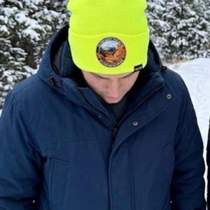 May include: A neon yellow beanie with a folded cuff and a circular patch on the front. The patch features a landscape illustration with text around the border. A small black rectangular tag is attached to the side of the cuff.