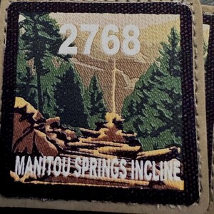 May include: A square fabric patch with a tan border and a dark brown edge. The center features a printed landscape of green pine trees and a rocky path. The white text 2768 is at the top, and the white text MANITOU SPRINGS INCLINE is at the bottom.