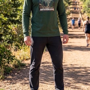 May include: A long-sleeve forest green crewneck shirt featuring a rectangular graphic print on the chest. The graphic depicts a mountain landscape with the text 2768 and MANITOU SPRINGS INCLINE. The shirt is paired with solid dark charcoal pants.