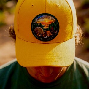 May include: A bright yellow baseball cap featuring a circular patch on the front. The patch displays a colorful landscape illustration of a mountain trail at sunset with the text The Manitou Springs Incline and Elevation 6530 / 2768 feet.