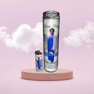 Peut inclure: Une bougie en verre blanc avec une photo d'une femme en robe bleue, avec le texte "Loving Mom" et "Linda Hen". Un briquet assorti plus petit est à côté de la bougie. La bougie et le briquet sont sur un socle en bois sur fond rose.