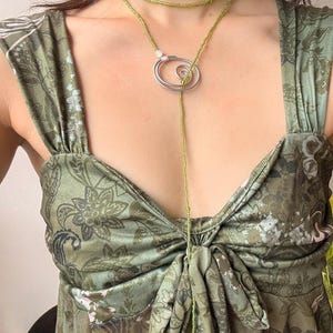 Puede incluir: Un vestido sin mangas en verde oliva con un estampado floral y de cachemira en verde oscuro, con un corpiño fruncido. Un collar largo de cuentas verde lima se envuelve alrededor del cuello, pasando a través de un colgante de espiral de metal plateado con una pequeña cuenta.
