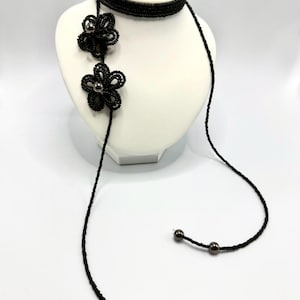 May include: A black beaded necklace with two flower pendants and dangling strands. The necklace is displayed on a white mannequin bust. The flower pendants have a dark center and are made of small, black beads. The necklace is a unique accessory.