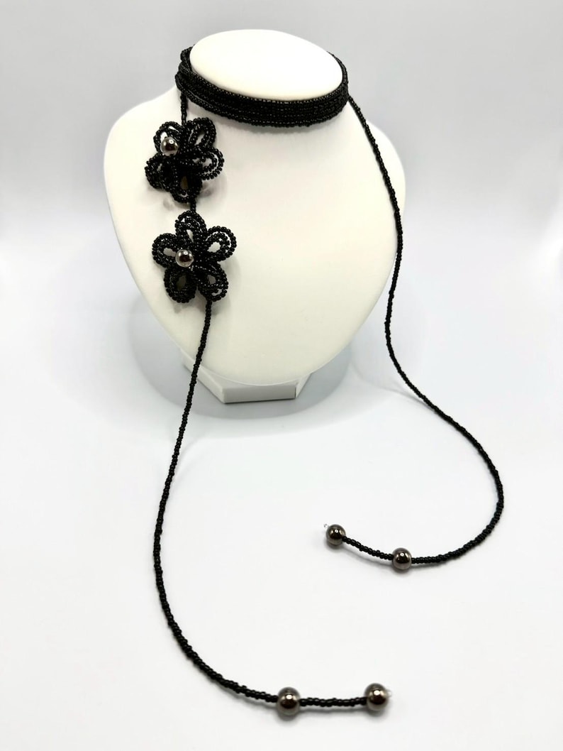 May include: A black beaded necklace with two flower pendants and dangling strands. The necklace is displayed on a white mannequin bust. The flower pendants have a dark center and are made of small, black beads. The necklace is a unique accessory.