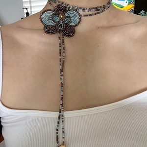 May include: A beaded choker necklace featuring a large flower pendant. The flower is brown with teal and gold accents. The necklace has a long beaded strand with a gold charm at the end. The choker is worn around the neck.