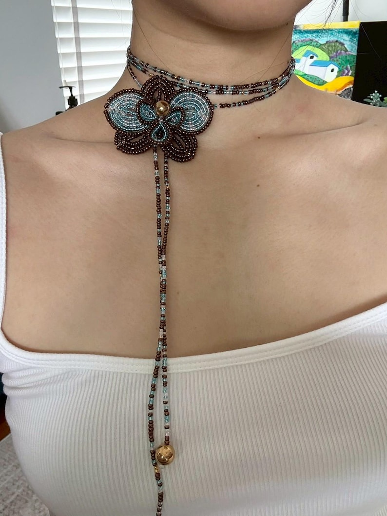 May include: A beaded choker necklace featuring a large flower pendant. The flower is brown with teal and gold accents. The necklace has a long beaded strand with a gold charm at the end. The choker is worn around the neck.