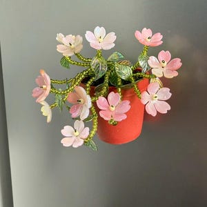 Puede incluir: Un imán decorativo con una planta en maceta en miniatura con flores rosas y blancas. Las flores tienen pétalos translúcidos y están unidas a tallos verdes con cuentas. La maceta es de color rojo vibrante.