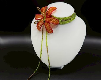 May include: A beaded choker necklace with a floral design. The necklace features a green beaded band and a flower with orange and red petals. The flower has long, beaded strands in green and brown. The necklace is displayed on a white mannequin.