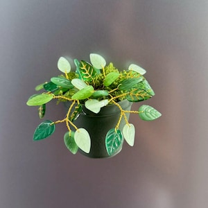 Könnte beinhalten: Ein kleiner, dunkelgrüner Keramiktopf mit einer Miniaturpflanze. Die Pflanze hat grüne Glasblätter und -stiele in verschiedenen Schattierungen mit goldenen Drahtakzenten. Die Blätter sind eine Mischung aus einfarbigen und gemusterten Designs.