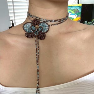 May include: A beaded choker necklace featuring a large, flower-shaped pendant. The pendant is brown and teal, with a gold center. The necklace has multiple strands of small beads in brown, teal, and gold.