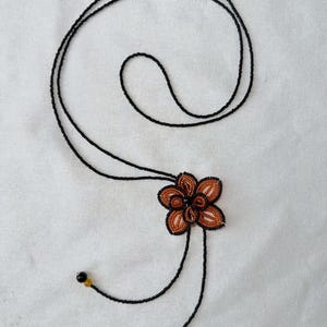 May include: A beaded necklace featuring a flower pendant. The flower is made of orange and black beads, with a dark center. The necklace has a long, black beaded chain that can be adjusted. The ends of the chain have small black and yellow beads.