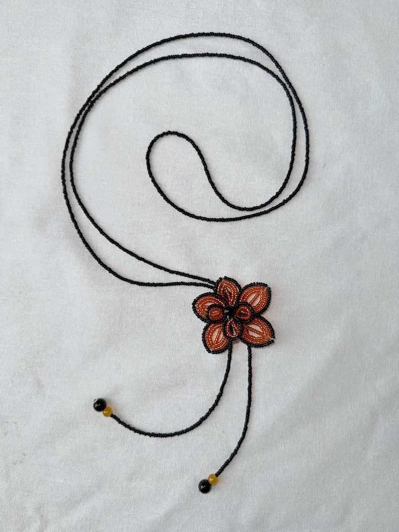 May include: A beaded necklace featuring a flower pendant. The flower is made of orange and black beads, with a dark center. The necklace has a long, black beaded chain that can be adjusted. The ends of the chain have small black and yellow beads.