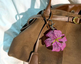 Könnte beinhalten: Eine braune Wildlederhandtasche mit Ledergriff und -besatz. Ein pinkfarbener und lila blütenförmiger Schlüsselanhänger hängt am Griff. Die Tasche hat einen Schnallenverschluss und ein kleines Lederetikett.