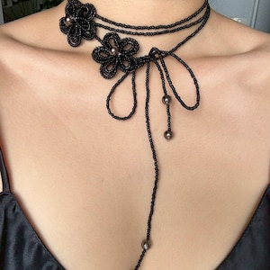 May include: A black beaded choker necklace with a floral design. The necklace features two beaded flower accents and a long, beaded strand with dangling beads. The necklace is worn on a person.