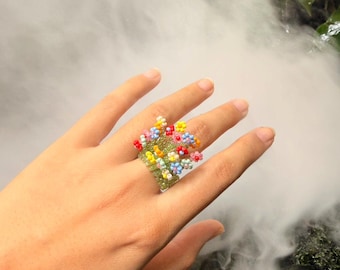 May include: A ring crafted from small green beads forming the band, topped with a cluster of tiny, multi-colored beaded flowers in shades of yellow, pink, blue, and orange. The floral arrangement sits prominently on the surface of the ring band.