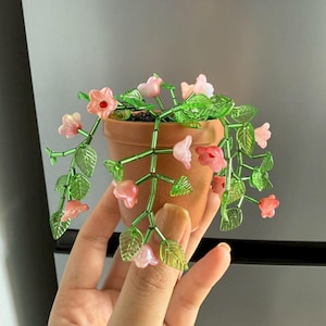 Może przedstawiać: Mały, terakotowy doniczka z rośliną z koralików. Roślina ma różowe kwiaty i zielone liście, a łodygi i liście są wykonane z zielonych koralików. Doniczka jest trzymana w dłoni.