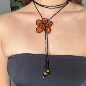 May include: A beaded necklace featuring a large orange flower pendant with black and yellow accents. The necklace has a black beaded choker and a long beaded strand. The flower pendant is the focal point of this handmade jewelry.