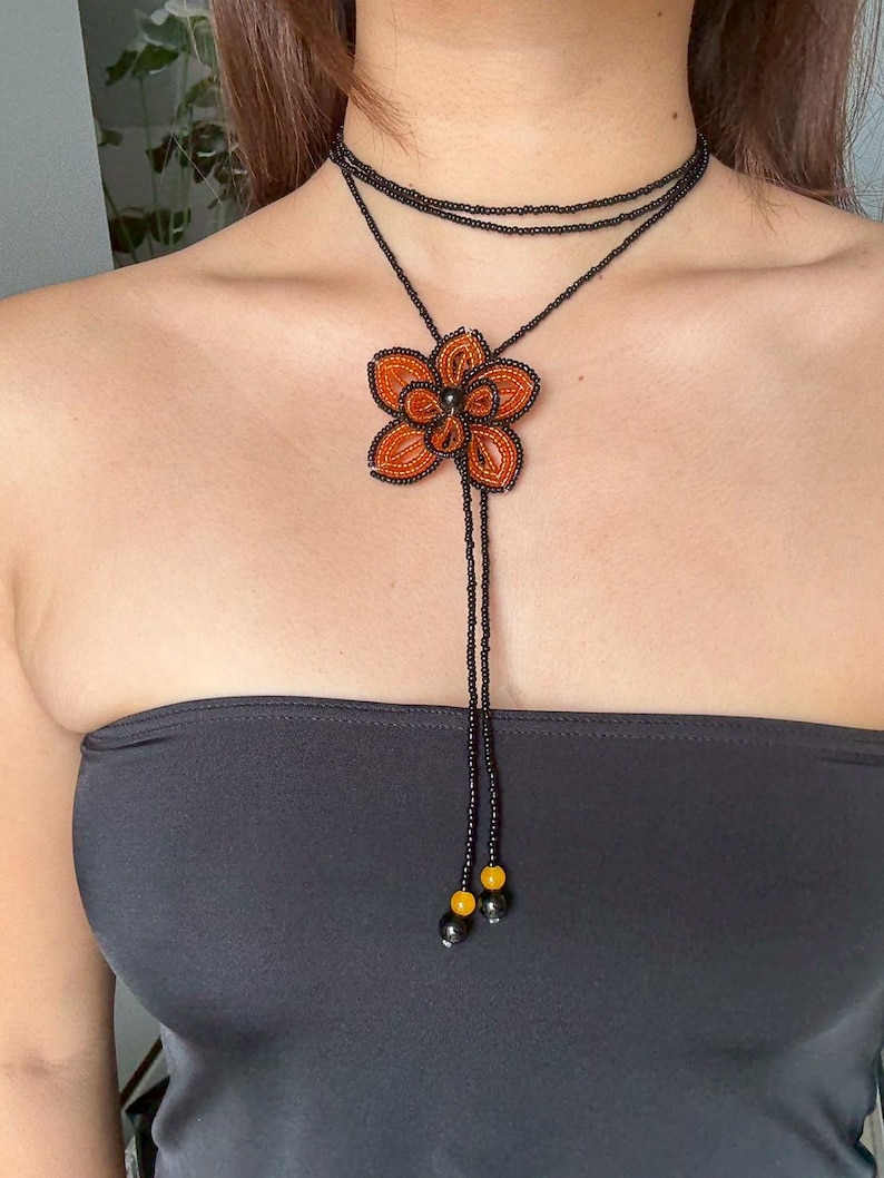 May include: A beaded necklace featuring a large orange flower pendant with black and yellow accents. The necklace has a black beaded choker and a long beaded strand. The flower pendant is the focal point of this handmade jewelry.