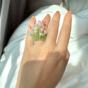 May include: A beaded ring featuring a cluster of colorful flowers. The ring has a green base with small, round beads forming the stems and leaves. The flowers are made of various colored beads, including red, pink, blue, and yellow.