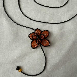 May include: A handmade necklace featuring a beaded flower pendant. The flower has orange and black petals with a black center. The necklace is made of black beads with yellow and black accents. The design is set against a white background.