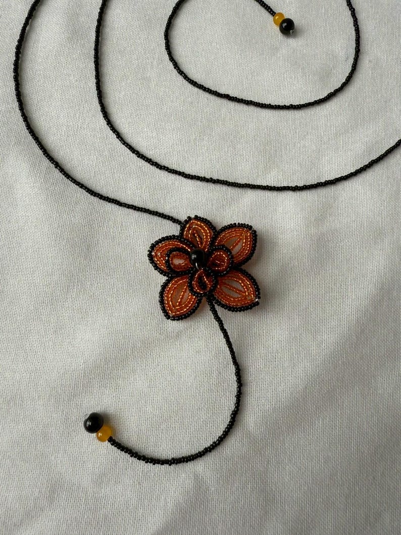 May include: A handmade necklace featuring a beaded flower pendant. The flower has orange and black petals with a black center. The necklace is made of black beads with yellow and black accents. The design is set against a white background.
