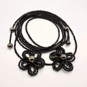 May include: Black beaded body chain featuring two flower designs with silver-colored centers. The chain is composed of multiple strands of small black beads, with silver beads at the ends. The flower designs are also made of black beads.