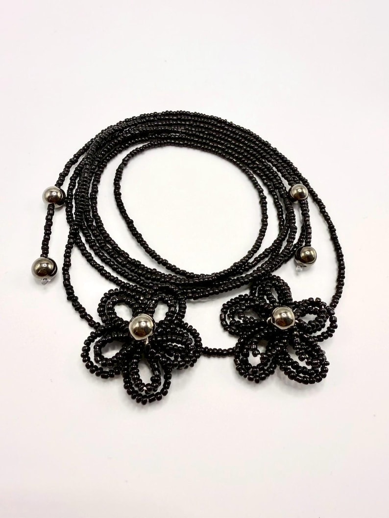 May include: Black beaded body chain featuring two flower designs with silver-colored centers. The chain is composed of multiple strands of small black beads, with silver beads at the ends. The flower designs are also made of black beads.