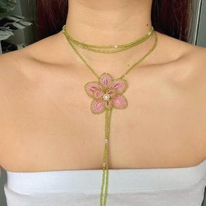 Puede incluir: Un collar de cuentas con un colgante de flor rosa y un centro de perla. El collar tiene un choker de cuentas verdes y una larga hebra de cuentas. El colgante de flores está detallado con cuentas rosas y doradas.