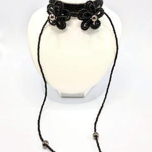 May include: Black beaded choker necklace with floral accents and dangling strands. The choker features two beaded flower designs with silver-colored centers. The necklace is displayed on a white mannequin head.