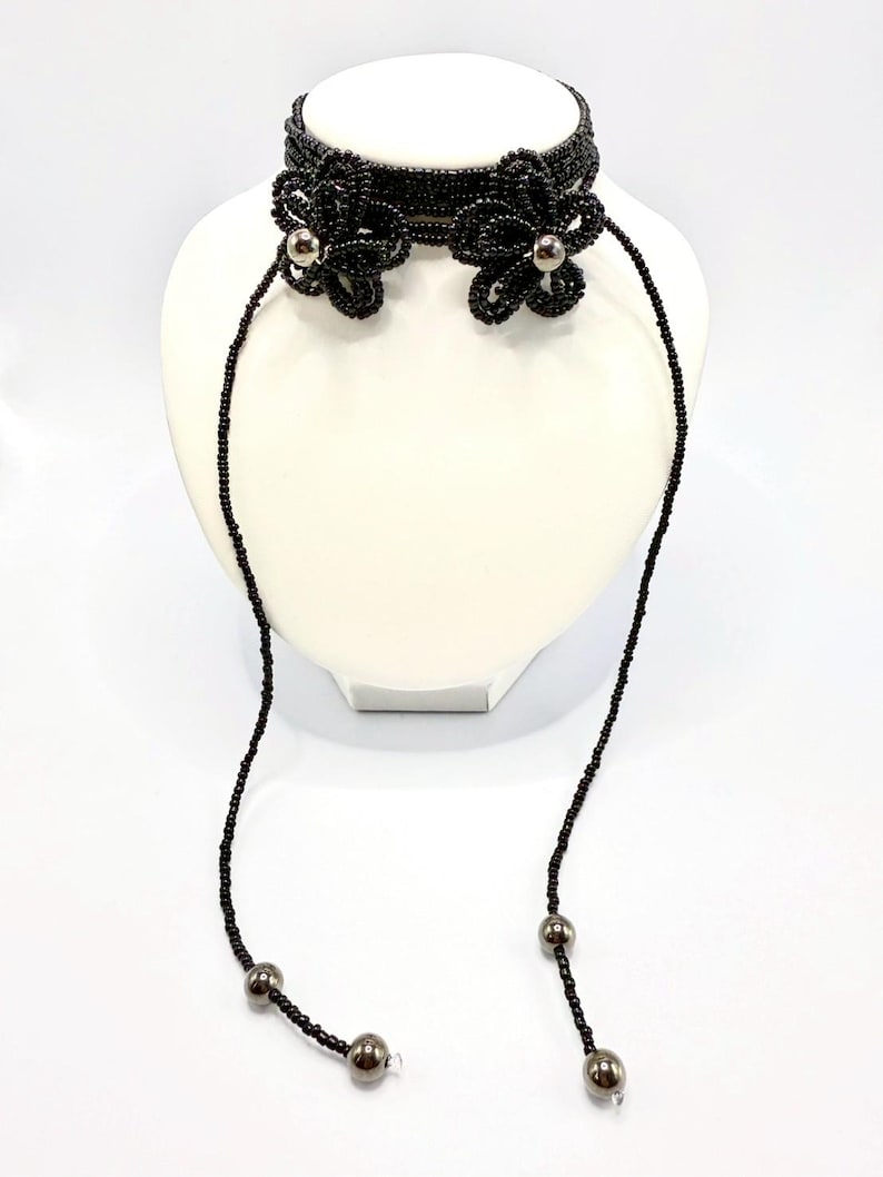 May include: Black beaded choker necklace with floral accents and dangling strands. The choker features two beaded flower designs with silver-colored centers. The necklace is displayed on a white mannequin head.