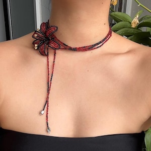 May include: A red and black beaded necklace with a floral design. The flower is the central feature, with gold-coloured accents. The necklace is a choker style with beaded strands that hang down, ending with silver beads.