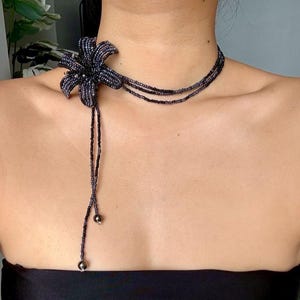 May include: A dark blue beaded necklace with a flower pendant. The flower is made up of many small beads. The necklace has a choker style with a beaded tassel. The beads are dark blue and black, creating a striking contrast.