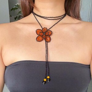 May include: A beaded necklace featuring a large orange flower pendant. The necklace has a black beaded choker and a long beaded strand with yellow and black accents. The flower pendant is intricately detailed with orange beads.