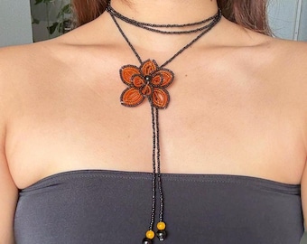May include: A beaded necklace featuring a large orange flower pendant. The necklace has a black beaded choker and a long beaded strand with yellow and black accents. The flower pendant is intricately detailed with orange beads.