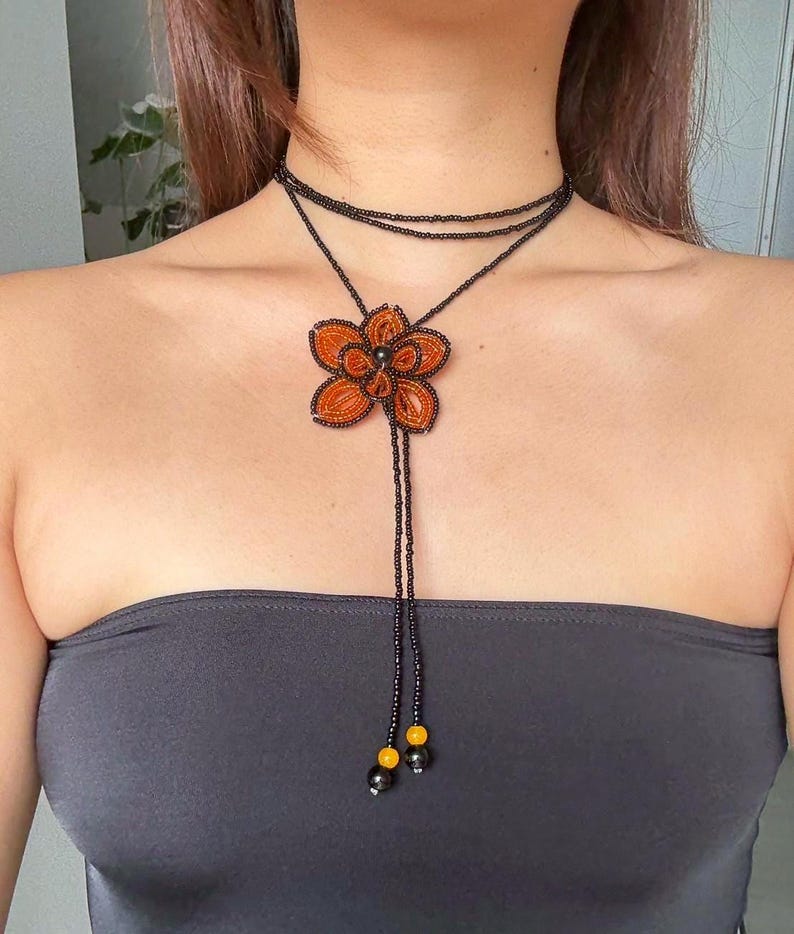 May include: A beaded necklace featuring a large orange flower pendant. The necklace has a black beaded choker and a long beaded strand with yellow and black accents. The flower pendant is intricately detailed with orange beads.