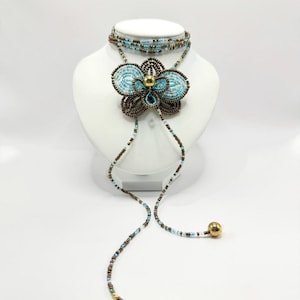 May include: A beaded necklace and choker set. The necklace features a large, flower-shaped pendant with turquoise and brown beads, accented by a gold-colored bead in the center. The choker is made of multiple strands of turquoise and brown beads. The necklace has two long beaded strands with gold-colored ball ends.